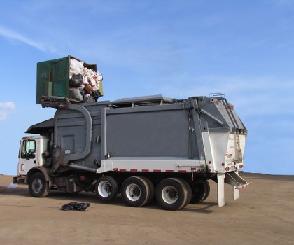 A large commercial waste collection truck parked on an open, paved area with a clear blue sky overhead. The truck has a modern, grey body designed for rubbish collection, with a side-loading mechanism visible, and a white cab at the front. The rear of the truck features a waste-compacting container with a closed, solid metal surface, along with a small tail lift or loading platform. In the foreground, there is a small pile of black waste bags on the ground. The background shows a flat, open landscape typical of outdoor rubbish removal sites or waste management depots. The image aligns with outdoor rubbish collection services provided by Commercial Waste Deptford, serving areas like Deptford and the surrounding postcode regions.