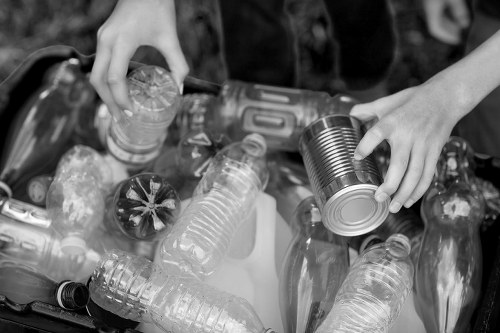 A close-up view of hands sorting and collecting empty plastic bottles and cans, including transparent water bottles and metal food tins, into a container or recycling bin. The bottles vary in size and shape, with some having ridged collars and others smooth surfaces, and are made of clear or metallic materials. The scene takes place outdoors on a paved surface, possibly in a garden or driveway, with a background of blurred foliage and natural elements. The person’s hands are actively handling the recyclable items, with visible fingers and part of the forearms, indicating an effort to separate waste for recycling or disposal. This image aligns with rubbish removal services provided by Commercial Waste Deptford in the area, emphasizing proper waste segregation and responsible disposal practices.